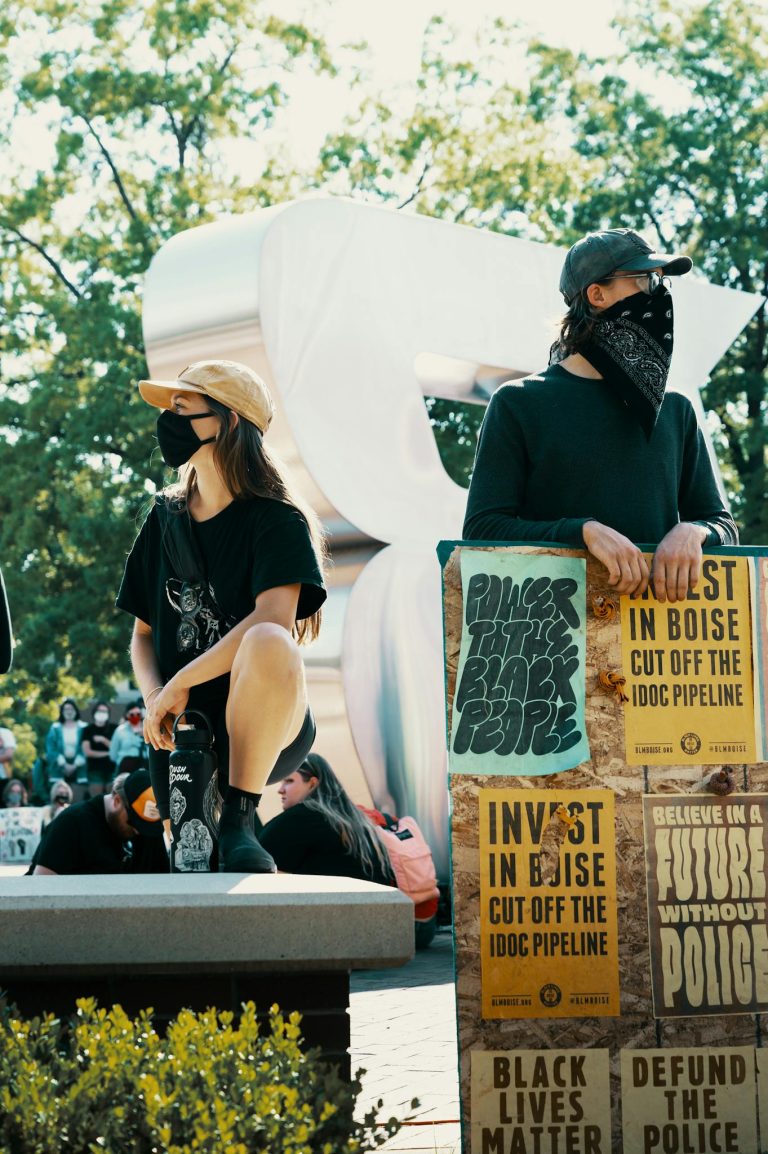 Protesters wearing masks hold signs demanding action at a city park during a sunny day demonstration.